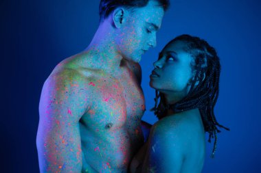 Renkli neon renkli vücut boyalı seksi çift mavi arka planda birbirlerine bakıyorlar. Cyan ışıklandırmalı, kaslı gövdeli üstsüz adam ve rasta saçlı çıplak Afrikalı Amerikalı kadın.