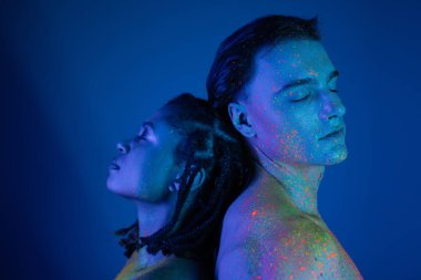 young interracial couple in colorful neon body paint standing back to back with closed eyes, african american woman with dreadlocks and man with bare shoulders on blue background with cyan lighting clipart