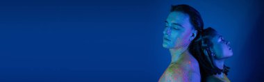 young interracial couple in colorful neon body paint, with bare shoulders and closed eyes, standing back to back on blue background with cyan lighting, banner clipart