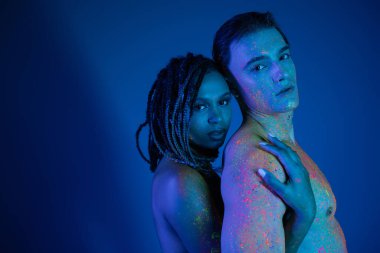 Rengarenk neon boyalı seksi bir çift mavi arka planda kameraya bakıyor. Cyan ışıklandırmalı, çıplak Afrikalı Amerikalı kadın kaslı vücutlu, üstsüz bir adamı kucaklıyor.