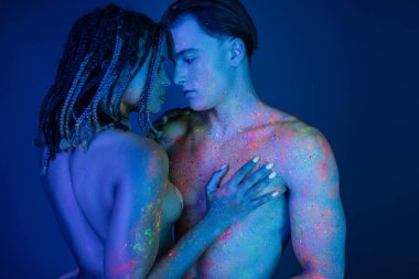 Rengarenk neon vücut boyalı ırklar arası bir çiftin mahrem anı. Çıplak Afrikalı Amerikalı kadın mavi arka planda çıplak kaslı adamın çıplak göğsüne dokunuyor.