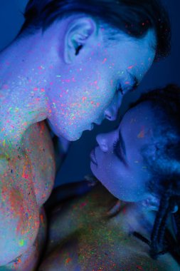 Renkli neon boyalı genç ve çok kültürlü bir çiftin mahrem anı. Çıplak göğüslü adam ve mavi arka planda seksi Afrikalı Amerikalı kadın.