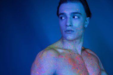 Genç, göz alıcı ve kaslı vücutlu, parlak ve renkli neon boyalı, siyan ışıklandırma efektiyle mavi arka plandan uzağa bakan kassız bir adam.