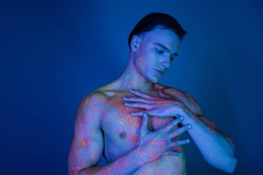 Çıplak göğsüne dokunan kaslı kassız genç adam mavi arka planda parlak ve renkli neon bir vücut boyası ve siyan ışık efektiyle poz veriyor.