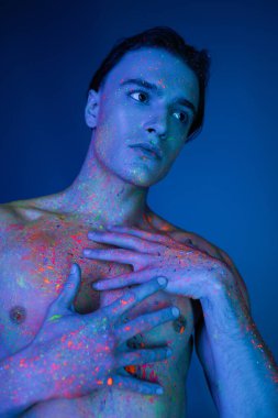 Işıl ışıl ve renkli neon vücut boyalarıyla poz veren yakışıklı ve atletik gömleksiz bir adam ve mavi arka planda siyan ışıklandırma efektiyle kaslı bir vücuda dokunuyor.