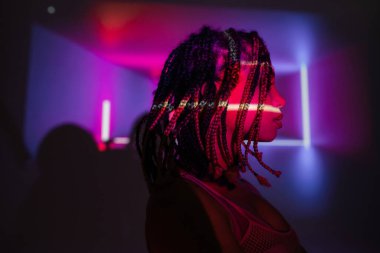 profile of young and charming african american woman with stylish dreadlocks standing on abstract purple background with vibrant neon rays and lighting effects clipart