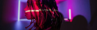 profile of young and captivating african american woman with dreadlocks posing on abstract purple background with radiant neon rays and lighting effects, banner clipart