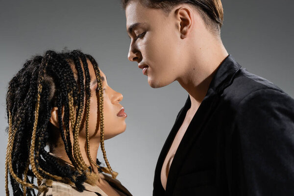 side view of sexy interracial couple, african american woman with stylish dreadlocks and young good looking man wearing black silk blazer on grey background