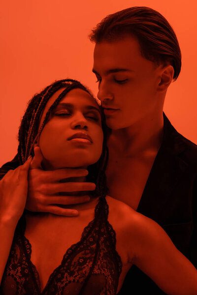 young and trendy man in black blazer hugging neck of captivating african american woman with dreadlocks and closed eyes, wearing lace lingerie on orange background with red lighting effect