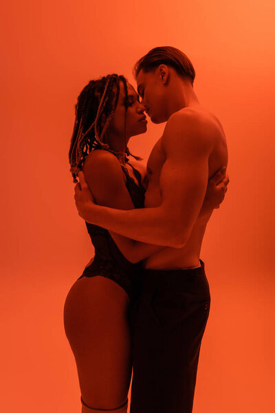 side view of interracial passionate couple, shirtless man in black pants with muscular torso and sexy african american woman in lace bodysuit embracing on orange background with red lighting effect