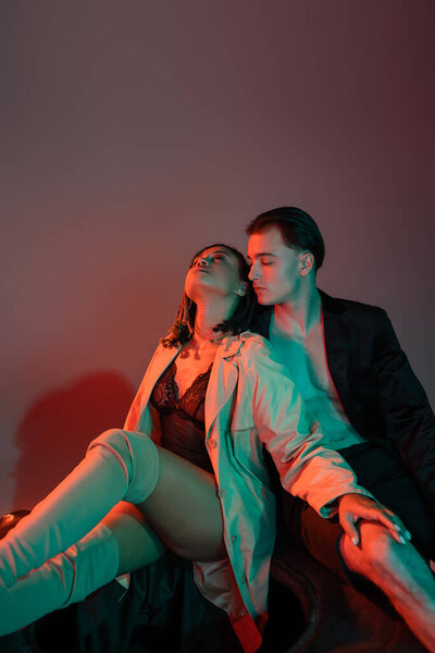 impassioned african american woman in black lace bodysuit and beige trench coat sitting near trendy man in black blazer on grey background with red lighting
