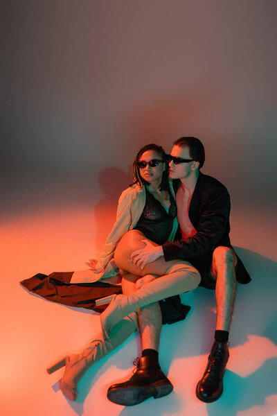 full length of glamour man in dark sunglasses and black blazer hugging leg of sassy african american woman in lace bodysuit, beige trench coat and over knee boots on grey with red lighting