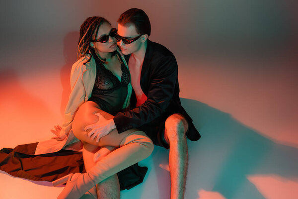 fashionable man in dark sunglasses and black blazer seducing african american woman in lace bodysuit and beige trench coat while hugging her leg on grey background with red lighting