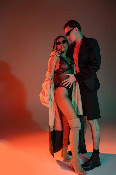 full length of stylish man in dark sunglasses and blazer hugging african american woman in black lace bodysuit, beige trench coat and over knee boots on grey background with red lighting