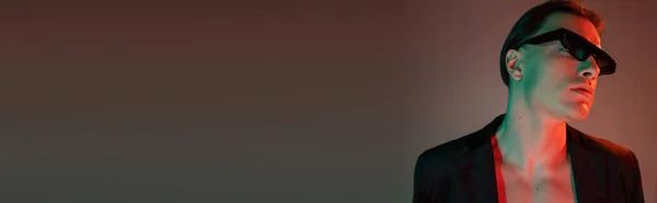 黒のブレザーと赤の照明とコピースペース バナーとグレーの背景に暗いトレンディーなサングラスのポーズでブルネットの髪を持つ魅力的な裸の胸の男の肖像画 — ストック写真