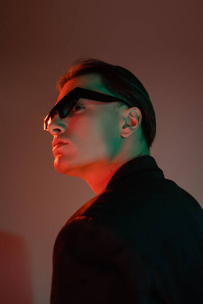 young and charismatic man with brunette hair posing in dark fashionable sunglasses and black blazer while looking away on grey background with red lighting