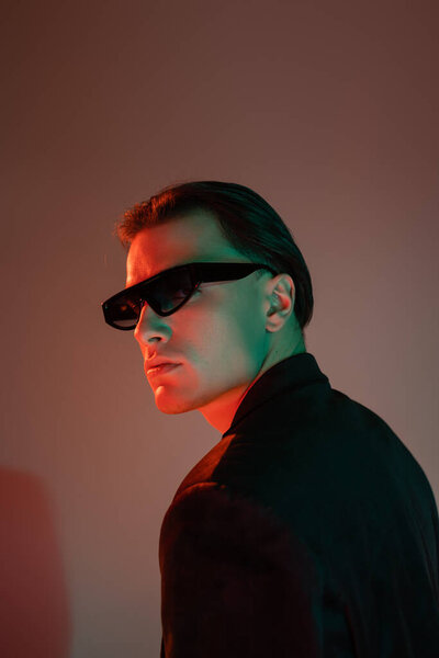 expressive and confident man with brunette hair, in dark trendy sunglasses and black blazer looking away while standing and posing on grey background with red lighting