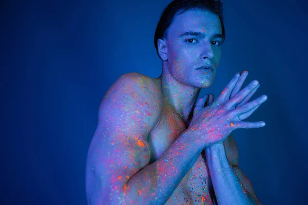 charismatic and self-assured shirtless man in radiant and multicolored neon body paint standing with joined hands and looking at camera on blue background with cyan lighting effect