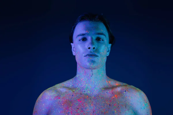 stock image portrait of charismatic man with naked shoulders posing in radiant and colorful neon body paint and looking at camera on blue background with cyan lighting effect