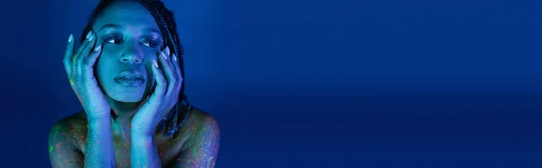 stock image portrait of youthful african american woman with dreadlocks, in colorful neon body paint, holding hands near face and looking away on blue background with cyan lighting effect, banner