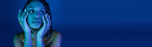 portrait of youthful african american woman with dreadlocks, in colorful neon body paint, holding hands near face and looking away on blue background with cyan lighting effect, banner