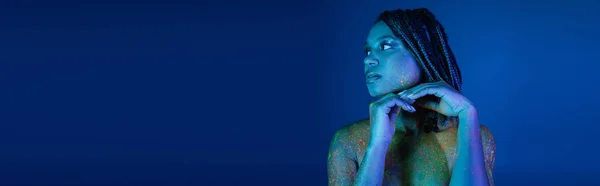 stock image sexy and eye-catching african american woman with dreadlocks, in multicolored neon body paint, holding hands near chin and looking away on blue background with cyan lighting effect, banner