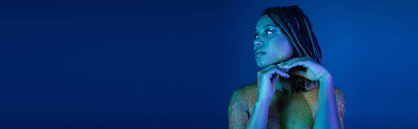 sexy and eye-catching african american woman with dreadlocks, in multicolored neon body paint, holding hands near chin and looking away on blue background with cyan lighting effect, banner