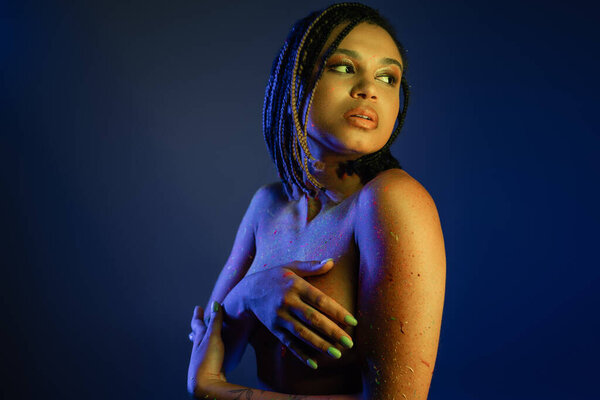 bare-chested african american woman with dreadlocks standing in colorful neon body paint, covering breast with hands and looking away on blue background with yellow lighting effect