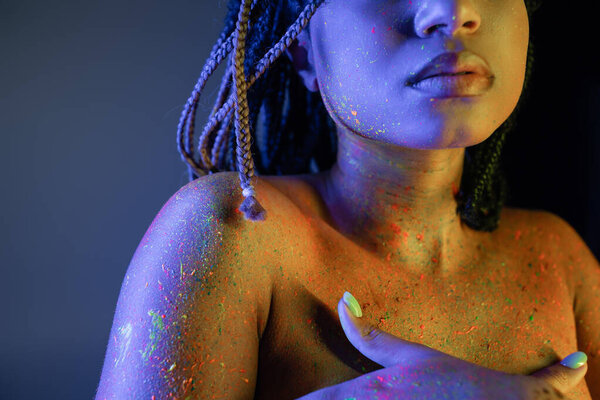 partial view of young and bare-chested african american woman in radiant and colorful neon body paint covering breast with hands on blue background with yellow lighting effect