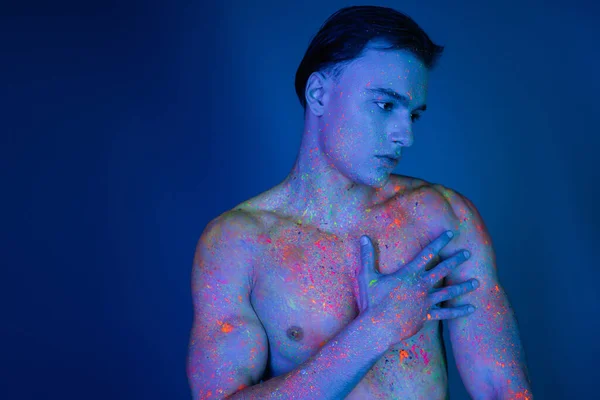 stock image handsome and young shirtless man with muscular torso, in colorful neon body paint, touching bare chest while standing on blue background with cyan lighting effect