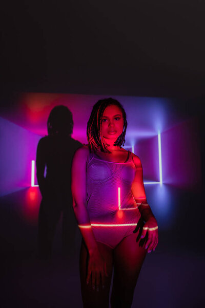 irresistible african american woman in net bodysuit, with stylish dreadlocks looking at camera while standing on abstract black and purple background with neon rays and lighting effects