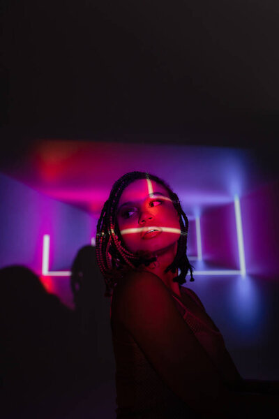 portrait of youthful and impassioned african american woman with dreadlocks looking away while standing on abstract black and purple background with neon rays and lighting effects