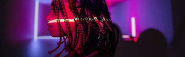 profile of young and captivating african american woman with dreadlocks posing on abstract purple background with radiant neon rays and lighting effects, banner