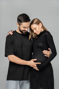 Siyah tişörtlü ve kotlu gülümseyen bir adam hamile ve şık bir kadının karnına dokunurken gri, büyüyen yeni bir hayat konseptinde ayakta durup sarılıyor.