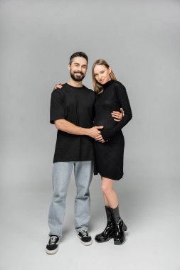 Full length of fashionable pregnant woman in black dress looking at camera and hugging bearded husband while standing together on grey background, growing new life concept clipart