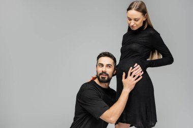 Excited bearded man in t-shirt touching and listening belly sounds of stylish pregnant wife in black dress while standing together isolated on grey, growing new life concept clipart