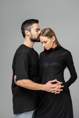 Tişörtlü sakallı koca, siyah elbiseli şık hamile kadının göbeğini öpüyor ve dokunuyor. Gri üzerinde tek başına duruyor. 