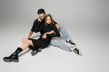 Smiling bearded man in jeans and t-shirt hugging fashionable pregnant woman in dress while sitting on grey background, new beginnings and parenting concept, husband and wife  clipart