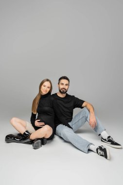 Smiling bearded man in t-shirt and jeans looking at camera while hugging stylish pregnant woman and sitting together on grey background, new beginnings and parenting concept, husband and wife  clipart