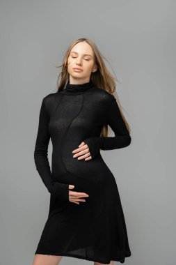 Relaxed fair haired and pregnant woman in stylish black dress closing eyes and touching belly while standing isolated on grey, new beginnings and maternity concept, mother-to-be clipart