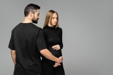 Siyah tişörtlü sakallı bir adam moda ve hamile bir kadının karnına dokunuyor. Gri üzerinde fotokopi alanı, yeni başlangıçlar ve beklenti konseptiyle tek başına dikilirken.  