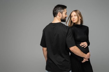Siyah tişörtlü, gülümseyen ve sakallı bir adam hamile bir kadının karnına dokunuyor ve gri, yeni başlangıçlar ve beklenti konseptinde tek başına dururken birbirlerine bakıyor.  