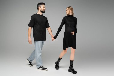 Full length of smiling man in jeans and black t-shirt holding hand of stylish pregnant wife in dress and walking on grey background, new beginnings and anticipation concept, expecting parents  clipart