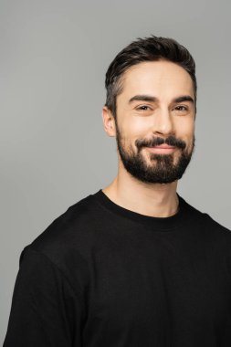 Portrait of positive bearded and brunette man in casual black t-shirt looking at camera while standing isolated on grey, masculine beauty concept, confident and charismatic  clipart