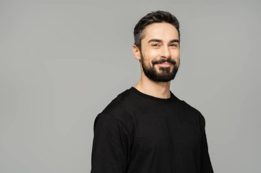 Portrait of cheerful brunette and bearded man in casual black t-shirt smiling at camera while standing isolated on grey, masculine beauty concept, confident and charismatic  clipart