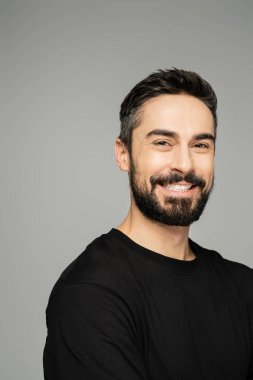 Portrait of confident and cheerful bearded man in casual black t-shirt looking at camera and standing isolated on grey, masculine beauty concept  clipart