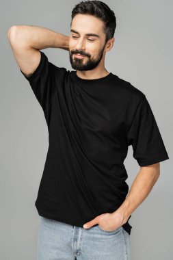 Portrait of relaxed and bearded man in casual black t-shirt and jeans holding hand in pocket and looking away while standing isolated on grey, masculine beauty concept  clipart