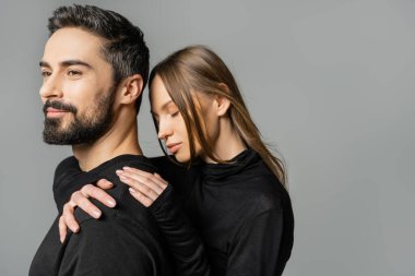 Siyah elbiseli, sırıtan ve sakallı kocasını kucaklayan açık renk saçlı bir kadın. Gri, karı-koca ilişkisi kavramında tek başına dikilirken.