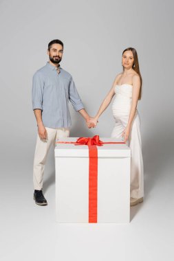 Smiling pregnant woman holding hand of husband and looking at camera while standing near big git box during gender reveal surprise party on grey background, expecting parents concept clipart