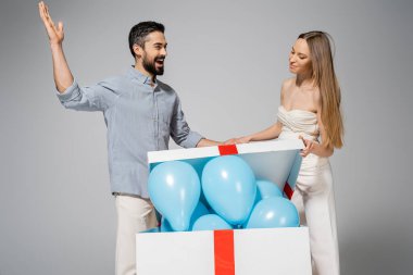 Excited bearded man looing at stylish pregnant wife opening big gift box with blue balloons during gender reveal surprise party and celebration isolated on grey, expecting parents concept, it`s a boy  clipart
